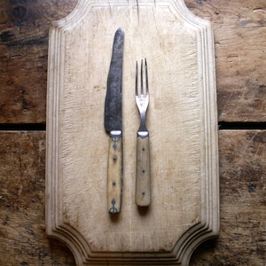 May include: A vintage wooden cutting board with a knife and fork. The board has a scalloped edge and is made of light wood. The knife and fork have bone handles and are made of metal.
