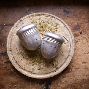 May include: Two silver tea infusers shaped like acorns, with small holes for brewing, rest on a wooden cutting board. Dried herbs and tea leaves are scattered around the infusers. The background is a rustic wooden surface.