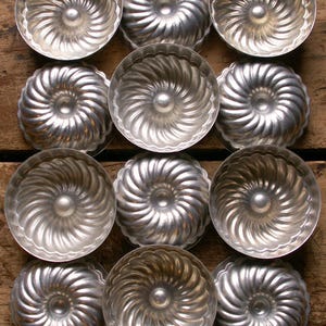 Op de afbeelding: Een verzameling van twaalf vintage, zilverkleurige metalen mallen, gerangschikt in een rasterpatroon. Elke mal heeft een spiraalvormig ontwerp en een centrale knop. De mallen worden waarschijnlijk gebruikt om te bakken of decoratieve voedingsmiddelen te creëren.