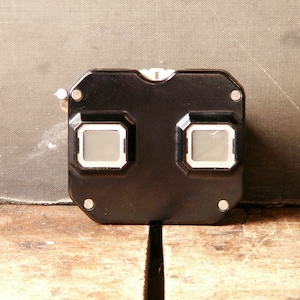 May include: A black plastic stereoscope viewer featuring two rectangular viewing lenses with white borders, a smooth glossy finish, and four small silver-toned metal rivets positioned at the corners of the front face of the device.
