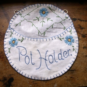 Puede incluir: Un agarrador blanco con texto bordado en azul que dice "Pot Holder". El agarrador tiene un borde cosido en azul y presenta flores azules bordadas con centros amarillos y hojas verdes. Hay un pequeño lazo blanco en la parte superior.