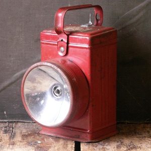 Könnte beinhalten: Eine rote Vintage-Metalllaterne mit einer großen kreisförmigen Linse und einem Griff. Die Laterne hat einen rechteckigen Körper und ein leicht abgenutztes Aussehen, was auf Alter und Gebrauch hindeutet. Die Linse ist reflektierend und der Griff ist gebogen.