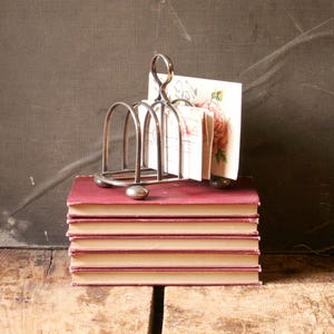 May include: A silver-toned metal letter holder with a looped handle, holding several postcards with floral designs. The holder sits atop a stack of five antique books with burgundy covers and cream-colored pages. The background is a dark gray.