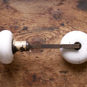 May include: A vintage door knob set featuring two white porcelain knobs connected by a metal rod. The knobs have a rounded shape and show signs of age, with some wear on the metal components. The set is placed on a wooden surface.