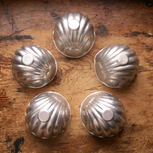 Könnte beinhalten: Fünf Vintage-Metallformen in Silber, jede in Form einer Jakobsmuschel, auf einer Holzoberfläche angeordnet. Jede Form hat ein kleines, kreisförmiges Detail oben. Die Formen dienen wahrscheinlich zum Backen oder zur Herstellung dekorativer Lebensmittel.