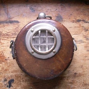 May include: A vintage brown leather diver's helmet-shaped clock. The clock face is visible through a circular, metal-rimmed window with a grid pattern. The clock has a metal loop at the top and a leather strap on the side.