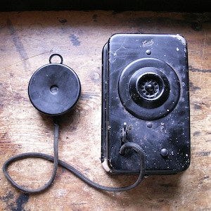 Puede incluir: Un teléfono de pared negro antiguo con un auricular circular y una base rectangular. El teléfono tiene un dial giratorio y un cable que conecta el auricular a la base. El teléfono está colocado sobre una superficie de madera.