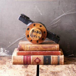 May include: A wooden object with black painted arms and a round center with the words "BABB CORES" painted on it. The object is sitting on top of a stack of three vintage books.