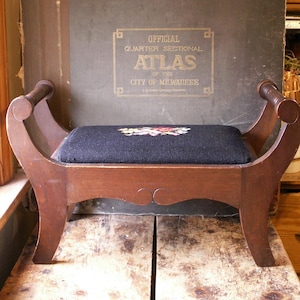Peut inclure: Un petit banc en bois avec une assise rembourrée bleu foncé ornée d'une broderie florale. Le banc a des pieds et des accoudoirs incurvés. Derrière le banc, un atlas vintage avec des lettres dorées.