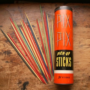 Può includere: Un set di gioco vintage Pick-Up Sticks. Il contenitore arancione e nero recita "PIX PIX PICK-UP STICKS" con "31 STICKS" in basso. I bastoncini sono di colore rosso, giallo, blu, verde e legno naturale.