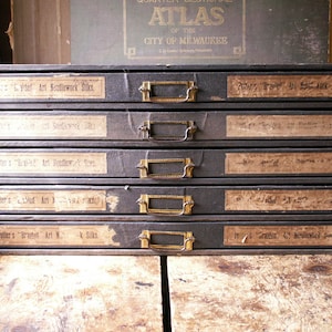 Puede incluir: Un mueble de almacenamiento vintage gris oscuro con cuatro cajones, cada uno etiquetado con etiquetas de papel envejecido y manijas de latón. Las etiquetas dicen "Art Needlework Silks" y otro texto relacionado. Un atlas desgastado es visible en el fondo.