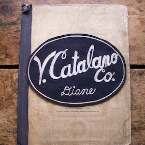 May include: An aged book with a dark blue spine and a black oval patch. The patch features the text "V. Catalano Co." in white script, with "Diane" below. The book's cover is beige with faded text and a decorative border.