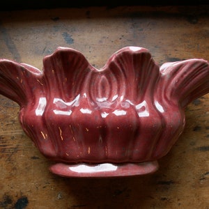 Puede incluir: Un cuenco de cerámica burdeos con un borde ondulado y una base de pedestal. El cuenco tiene un acabado brillante. El diseño recuerda a una flor o una concha. El cuenco es probablemente para fines decorativos o de servicio.