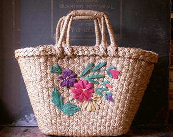 Könnte beinhalten: Eine gewebte Stroh-Tragetasche mit rechteckiger Form und geflochtenen Griffen. Die Tasche ist mit bunten Stoffblumen in Rosa-, Lila- und Gelbtönen mit grünen Blättern verziert. Die Tasche steht auf einer Holzoberfläche.