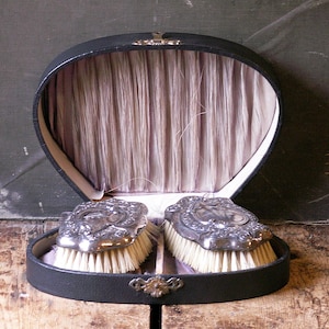 Puede incluir: Un antiguo neceser ovalado negro con dos cepillos plateados. Los cepillos tienen cerdas blancas y detalles ornamentados. El conjunto está forrado con una tela rosa claro y se muestra abierto, revelando el contenido.