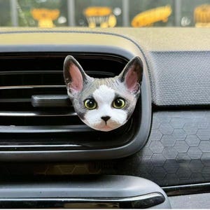 May include: A cat-shaped car air vent clip. The cat has gray and white fur, with large green eyes and pink ears. The clip is inserted into a black car vent, with a textured dashboard in the background.