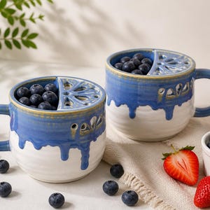 May include: Two ceramic mugs with a blue and white glaze, filled with blueberries. The mugs have a unique design with a lemon slice-shaped opening. Strawberries and blueberries are scattered around the mugs, creating a fresh, inviting scene.
