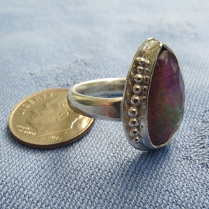 May include: A silver ring with a large, pear-shaped gemstone. The gemstone is a deep purple color with green and gold accents. The ring is set with a beaded band.