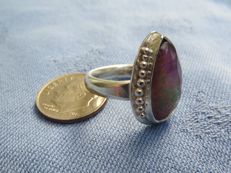 May include: A silver ring with a large, pear-shaped gemstone. The gemstone is a deep purple color with green and gold accents. The ring is set with a beaded band.