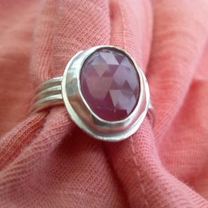 May include: A silver ring with a large, faceted, oval-shaped, ruby-colored gemstone. The ring has a simple band with three parallel lines. The gemstone is set in a silver bezel, and the ring is displayed on a pink fabric.