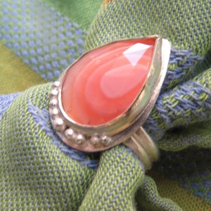 May include: A silver ring with a teardrop-shaped pink agate stone set in a silver bezel. The ring has a beaded edge and a smooth, polished finish.