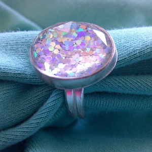 May include: A silver ring with a round iridescent glitter top. The glitter is a mix of white, pink, and purple.
