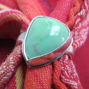 May include: A silver ring with a large, light green, triangular stone. The ring is set against a red and orange patterned fabric background. The stone has a smooth, polished surface.