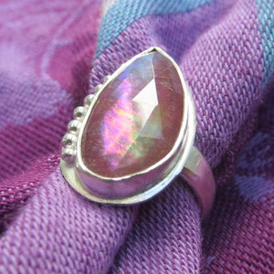 May include: A silver ring with a pear-shaped pink gemstone set in a bezel setting. The ring has a beaded detail around the setting.