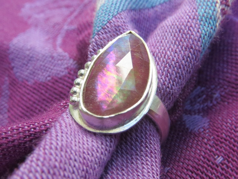 May include: A silver ring with a pear-shaped pink gemstone set in a bezel setting. The ring has a beaded detail around the setting.