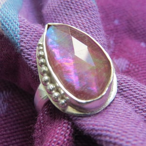 May include: A pearlescent pink gemstone ring with a silver setting and a beaded border.