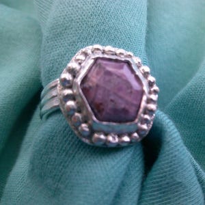 May include: A silver ring with a large, hexagonal, purple gemstone. The gemstone is set in a silver bezel surrounded by small silver beads. The ring is on a teal fabric background.