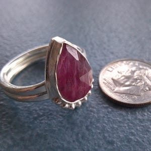May include: A silver ring with a pear-shaped pink gemstone set in a bezel setting. The ring is double-banded and has a textured finish.