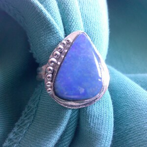 May include: A silver ring with a large, pear-shaped blue opal gemstone. The ring has a textured silver band with small beaded accents along the side of the opal. The ring is set against a light blue fabric.