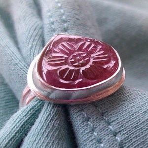 May include: A silver ring with a pink carved flower gemstone. The ring is on a blue fabric background.