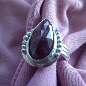 May include: A silver ring with a large, faceted, teardrop-shaped, dark red gemstone. The ring has a decorative beaded border around the stone and a simple band with three parallel lines. The ring is set against a soft, pinkish-purple fabric.