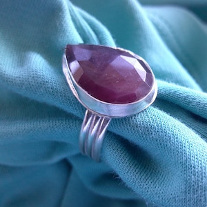 May include: A silver ring with a large, pear-shaped, faceted, dark purple gemstone. The gemstone is set in a silver bezel, and the ring has a triple-banded shank. The ring is displayed on a blue fabric background.
