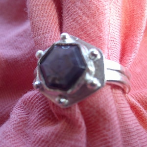 May include: A silver ring with a dark purple, hexagonal gemstone. The ring has a unique design with small, round embellishments around the gemstone and a double band. The ring is displayed on a pink fabric.
