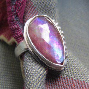 May include: A silver ring with a large, pear-shaped, iridescent gemstone set in a bezel setting. The gemstone is a deep pink with blue and purple iridescence. The ring band is decorated with small beads.