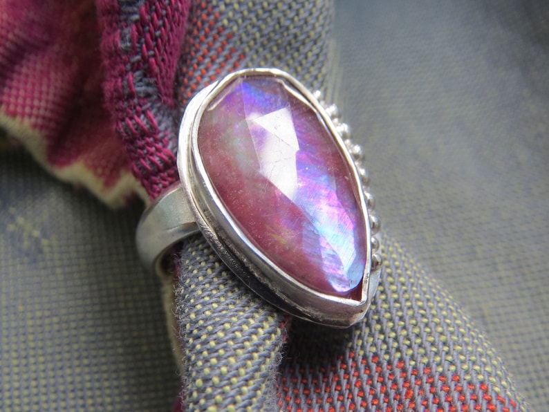 May include: A silver ring with a large, pear-shaped, iridescent gemstone set in a bezel setting. The gemstone is a deep pink with blue and purple iridescence. The ring band is decorated with small beads.