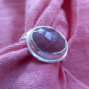 May include: A silver ring with a large, faceted, oval-shaped, pink gemstone. The ring is set against a pink fabric background, creating a contrast between the cool metal and the warm color of the fabric.