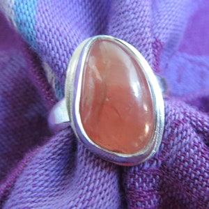 May include: A silver ring featuring a smooth, polished, teardrop-shaped peach-colored gemstone set in a simple metal bezel. The ring is positioned against a textured purple fabric background with visible woven threads and subtle blue highlights.
