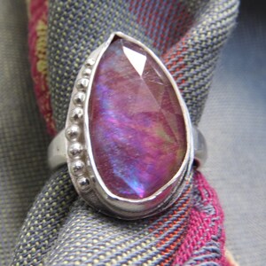 May include: A silver ring with a large, iridescent purple gemstone set in a bezel setting. The ring has a beaded band.