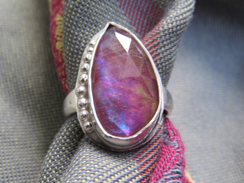 May include: A silver ring with a large, iridescent purple gemstone set in a bezel setting. The ring has a beaded band.