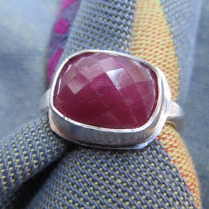 May include: A silver ring with a large, faceted, red gemstone. The ring is set on a fabric background.