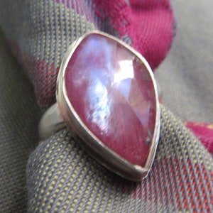 May include: A silver ring featuring a large, pear-shaped, faceted gemstone with a deep pink hue and iridescent highlights. The ring is set against a backdrop of a patterned fabric in shades of gray, red, and pink.