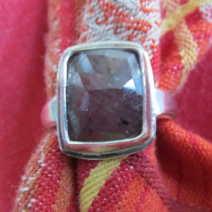 May include: A silver ring with a large, dark, rectangular gemstone. The gemstone has a faceted surface and a dark, brownish-purple color. The ring is set on a red and orange patterned fabric.