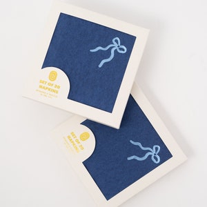 May include: Two boxes of blue napkins with a light blue ribbon design. The boxes are labelled "Set of 20 Napkins" and "Hand-Made & Printed in the USA".