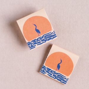 May include: Two matchboxes with a sunset design. Each box features an orange sun, blue water, and a blue heron silhouette. The boxes are rectangular with a cream-colored background.