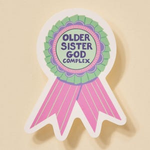 Op de afbeelding: Een sticker in de vorm van een lint met de tekst "OLDER SISTER GOD COMPLEX". De sticker heeft een groen en paars cirkelvormig ontwerp met roze linten.