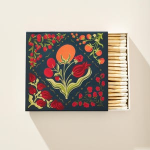 May include: A square matchbox with a dark blue cover featuring a botanical illustration of red berries, green leaves, and an orange fruit. The box is partially open, revealing a row of wooden matches with white tips arranged in a neat vertical line.